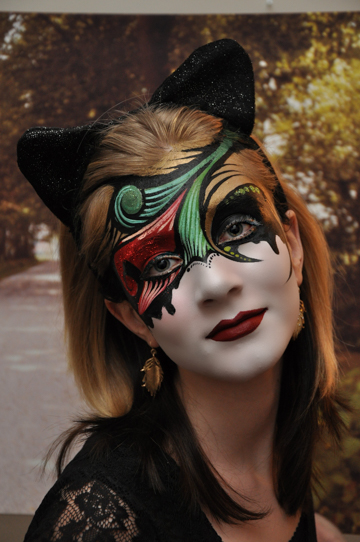 red haird woman with black cat ears on her head and a colorful mask painted on her face with red and green accents